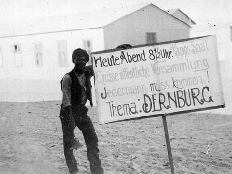 Mann mit handgemaltem Schild, das zu einer Versammlung zum Thema Dernburg einlädt